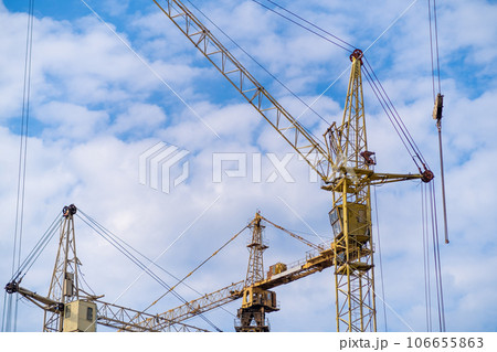A crane and a building under construction against a blue sky background. Builders work on large construction sites, and there are many cranes working in the field of new construction. A crane and a building under construction against a blue sky background. Builders work on large construction sites, and there are many cranes working in the field of new construction. 106655863