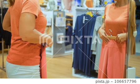 Trendy girl looks in mirror and try outfits in modern shopping centre department store. Happy shopper girl posing in mirror in shopping mall. Sporty girl customer is looking and buying sport clothes. 106657796