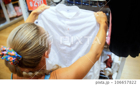 Young female shopper examines clothes from rack in mall store.Portrait of shopaholic woman shopping in fashion boutique. Selects touches clothes sale on Black Friday attractive girl in clothing store. 106657822
