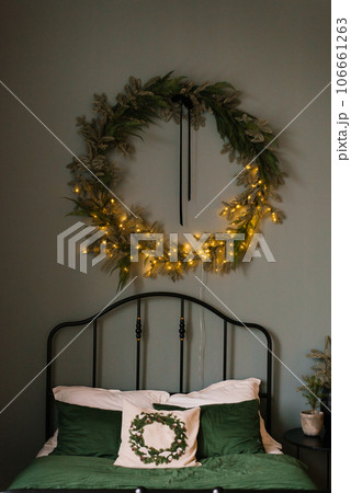 Scandinavian bedroom with white and green bed linen and Christmas decorations made of fir branches, garlands and wreaths on the wall Scandinavian bedroom with white and green bed linen and Christmas decorations made of fir branches, garlands and wreaths on the wall 106661263