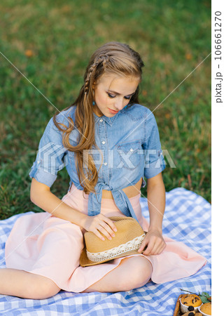 Young woman is sitting on a picnic blanket and holding a straw hat in her hands Young woman is sitting on a picnic blanket and holding a straw hat in her hands 106661270