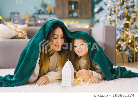 Mom and daughter are lying under a blanket and looking at a glowing toy house, waiting for a Christmas miracle. The concept of a children's holiday 106661274