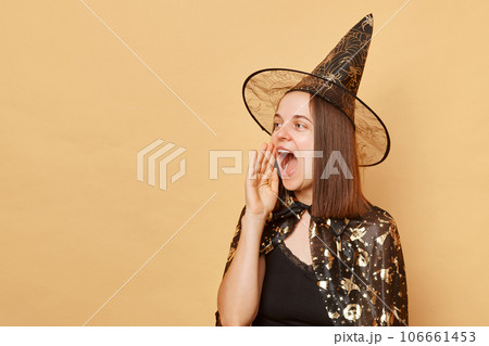 Cheerful young girl wearing black halloween garment and cone posing isolated over beige background keeps hand near mouth screaming making announcement 106661453