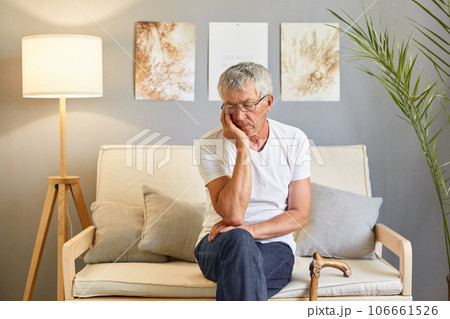 Elderly man with a cane. Grandfather's old on couch. Bored unhappy man pensioner wearing white T-shirt and jeans sitting on sofa in living room holding walking cane Elderly man with a cane. Grandfather's old on couch. Bored unhappy man pensioner wearing white T-shirt and jeans sitting on sofa in living room holding walking cane 106661526