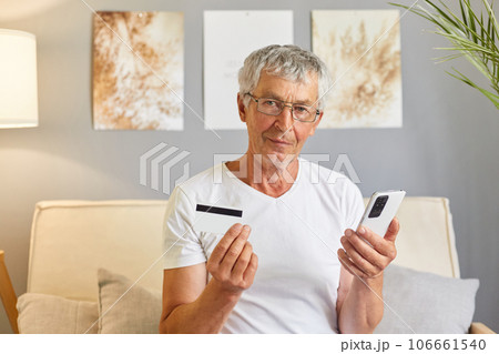 Calm mature man using mobile phone and making online payment by credit card, customer shopping from home, paying for purchase on Internet stores sitting on sofa at home interior. Calm mature man using mobile phone and making online payment by credit card, customer shopping from home, paying for purchase on Internet stores sitting on sofa at home interior. 106661540