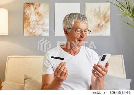 Focused senior man using smartphone and making online payment by credit card using banking or ecommerce app for transactions sitting on couch in living room. Focused senior man using smartphone and making online payment by credit card using banking or ecommerce app for transactions sitting on couch in living room. 106661541