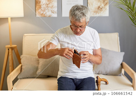 Sad unhappy senior man looking inside empty wallet looking for money wearing white T-shirt sitting on cough in living room has not money to buy what he needs. Sad unhappy senior man looking inside empty wallet looking for money wearing white T-shirt sitting on cough in living room has not money to buy what he needs. 106661542