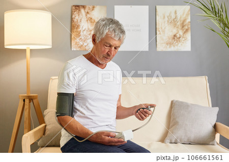 Mature gray haired male patient wearing white T-shirt and jeans using modern tonometer at home, making medicine self checkup man measuring his blood pressure while sitting on sofa in living room. Mature gray haired male patient wearing white T-shirt and jeans using modern tonometer at home, making medicine self checkup man measuring his blood pressure while sitting on sofa in living room. 106661561