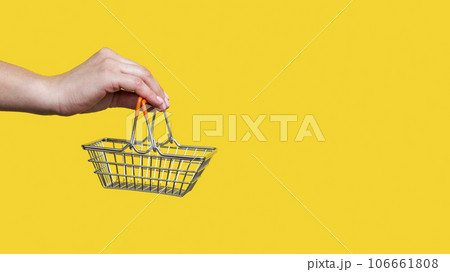 Female hand holds toy metal shopping basket on yellow background. Female hand holds toy metal shopping basket on yellow background. 106661808