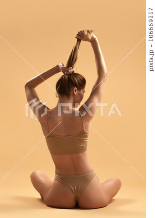 Rear view of young girl with slim body, buttocks sitting in lotus pose in beige underwear against sand studio background. healthy back. 106669117