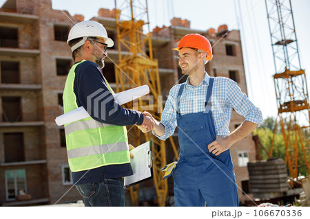 Worker and architect watching some details on a construction. Two engineers talking at building site with construction structure in background. Men wearing helmets. Worker and architect watching some details on a construction. Two engineers talking at building site with construction structure in background. Men wearing helmets. 106670336