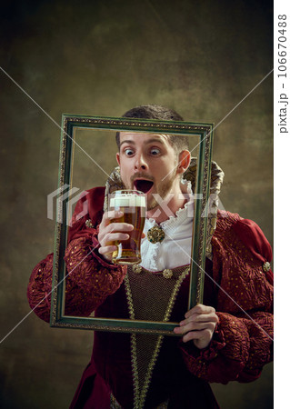 Medieval royal person, prince in female dress holding picture frame and drinking beer against dark green background Medieval royal person, prince in female dress holding picture frame and drinking beer against dark green background 106670488