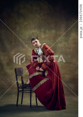 Young medieval prince, royal person leaning on chair, wearing female dress against dark green background. Renaissance style 106670496