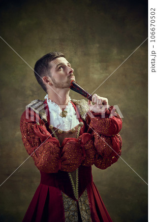 Thoughtful young man, medieval prince in female dress with fan against vintage green background. Equality Thoughtful young man, medieval prince in female dress with fan against vintage green background. Equality 106670502