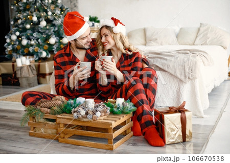 Romantic couple celebrating Christmas on a floor near Christmas tree. Blonde woman and brunette man wearing plaid pajamas. Romantic couple drinking a tea from a mugs. Romantic couple celebrating Christmas on a floor near Christmas tree. Blonde woman and brunette man wearing plaid pajamas. Romantic couple drinking a tea from a mugs. 106670538