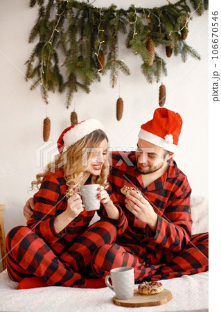 Lovely couple celebrating christmas on a bed. Blonde woman and brunette man wearing plaid pajamas. Romantic couple drinking a tea from a mug and eating a donuts. 106670546