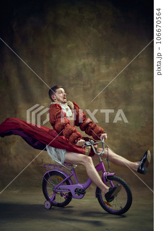 Young man, medieval royal person in female dress riding little bike against vintage background. Humorous emotions. Speed 106670564