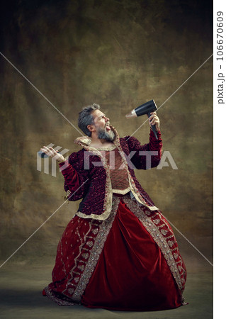 Gray-haired king, monarch in female dress singing in hair dryer while doing hairstyle against vintage background 106670609