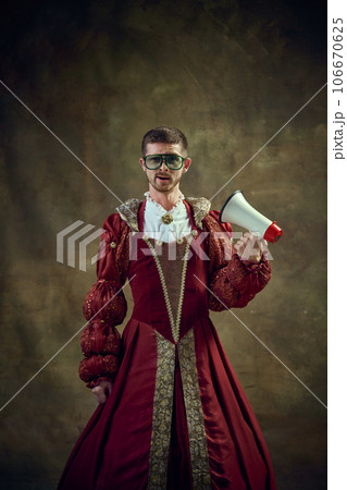 Young guy in image of medieval royal person wearing female dress and sunglasses standing with megaphone against vintage background Young guy in image of medieval royal person wearing female dress and sunglasses standing with megaphone against vintage background 106670625