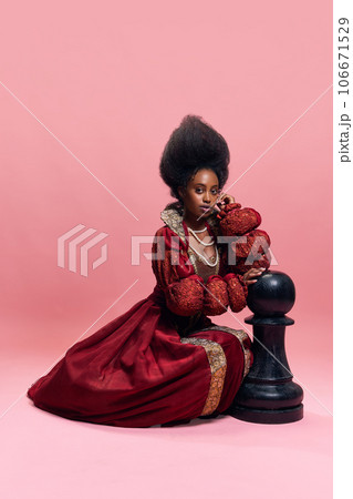 Checkmate. Portrait of medieval princess, queen in red costume playing with giant chess piece against pink studio background 106671529