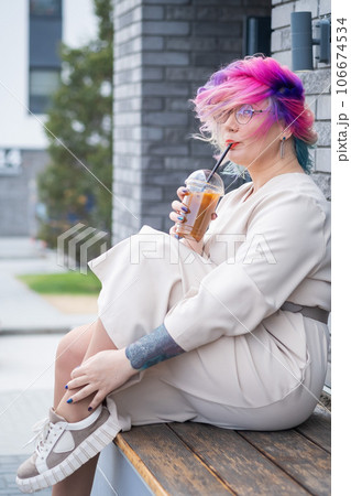 Close-up portrait of curly Caucasian woman with multi-colored hair wearing glasses. The hairstyle model is drinking a cold drink 106674534