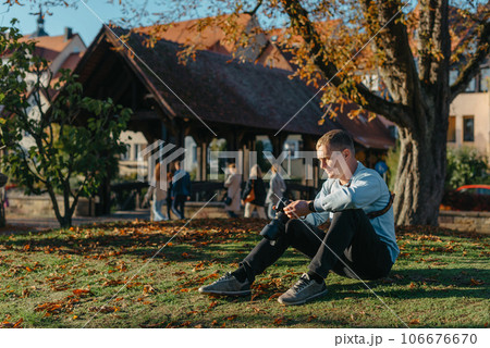 Professional Photographer Taking Picture Of Beautiful Autumn Park. Man Professional Photographer Sit With Camera And With Smartphone In Autumn Park. Retouched, Vibrant Colors, Brownish Tones. 106676670