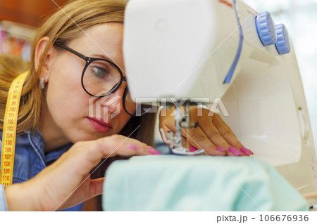 Woman seamstress in glasses works on sewing-machine at her workplace in workshop Woman seamstress in glasses works on sewing-machine at her workplace in workshop 106676936