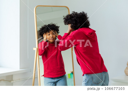 Love yourself. Beautiful young smiling african american woman dancing touching curly hair enjoying her mirror reflection. Black lady looking at mirror looking confident and happy. Self love concept 106677103