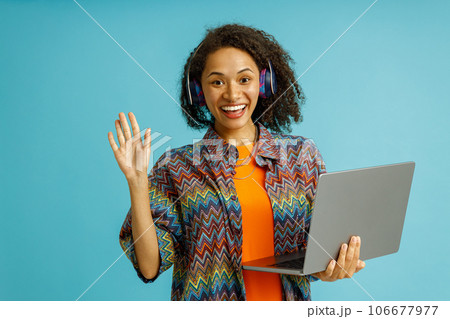 Smiling woman in headphones holding laptop and waving hi in camera on blue studio background 106677977