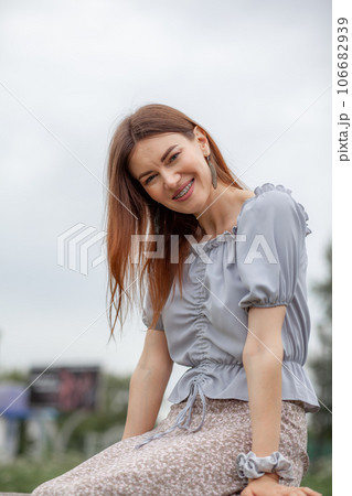 Smile of a young and beautiful girl with braces on her white teeth. A girl with long hair and a dress poses in the park. Straightening crooked teeth using a braces system. 106682939