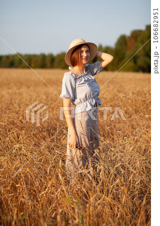 A young beautiful girl with braces on her teeth and long hair poses in a wheat field in the summer at sunset. The girl holds a hat in her hand against the background of a wheat field. 106682951