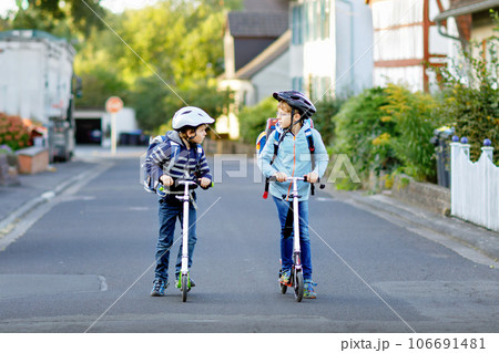 Two school kid boys in safety helmet riding with scooter in the city with backpack on sunny day. Happy children in colorful clothes biking on way to school. Two school kid boys in safety helmet riding with scooter in the city with backpack on sunny day. Happy children in colorful clothes biking on way to school. 106691481