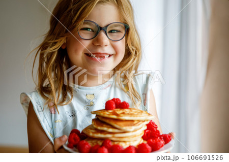 Little happy preschool girl with a large stack of pancakes and raspberries for breakfast. Positive child eating healthy homemade food in the morning. 106691526