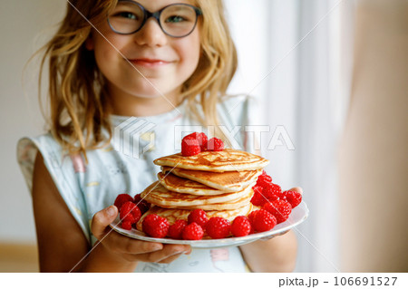 Little happy preschool girl with a large stack of pancakes and raspberries for breakfast. Positive child eating healthy homemade food in the morning. 106691527