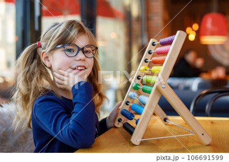 Little preschool girl playing with educational wooden rainbow toy counter abacus. Healthy happy child with glasses learning to count and colors, indoors on sunny day. Little preschool girl playing with educational wooden rainbow toy counter abacus. Healthy happy child with glasses learning to count and colors, indoors on sunny day. 106691599