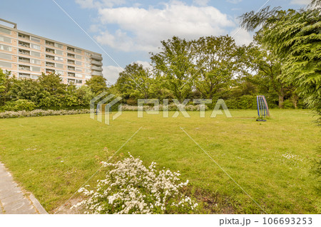 a grassy area in front of an apartment building with white flowers on the grass and trees around it, surrounded by blue sky 106693253