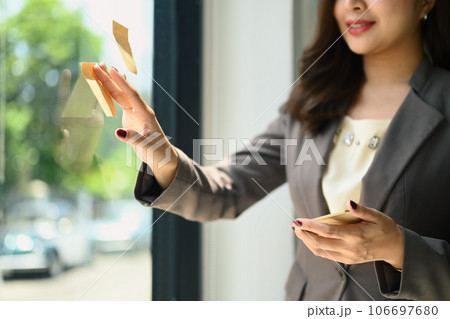 Smiling businesswoman sticking adhesive notes on glass window, planning a project and sharing ideas 106697680