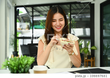 Pretty asian female worker sitting in office desk and eating sandwich for lunch. People and food concept 106697681