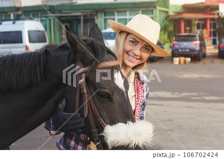 Young woman in straw hat standing behind dark brown horse, smiling, detail on their faces, city buildings in background 106702424