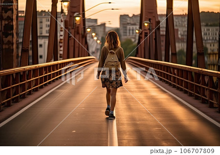 a woman walking across a bridge in the middle of the image is an orange sky and there are buildings behind her 106707809