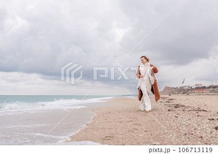 Blond woman Christmas tree sea. Christmas portrait of a happy woman walking along the beach and holding a Christmas tree on her shoulder. She is wearing a brown coat and a white suit. 106713119