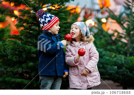 Two little smiling kids, boy and girl eating crystalized sugared apple on German Christmas market. Happy friends in winter clothes with lights on background. Family, tradition, holiday concept Two little smiling kids, boy and girl eating crystalized sugared apple on German Christmas market. Happy friends in winter clothes with lights on background. Family, tradition, holiday concept 106718688