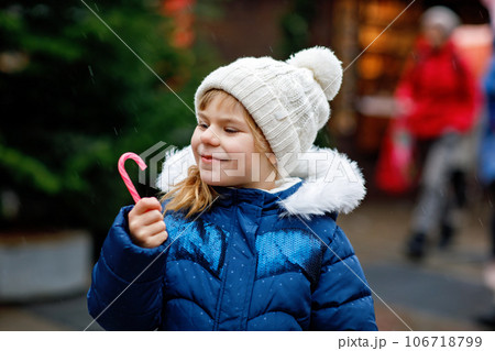 Little cute preschool girl with candy cane from a sweets stand on Christmas market. Happy child on traditional family market in Germany. Preschooler in colorful winter clothes during snowfall 106718799