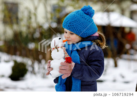 Cute little toddler girl making mini snowman and eating carrot nose. Adorable healthy happy child playing and having fun with snow, outdoors on cold day. Active leisure with children in winter Cute little toddler girl making mini snowman and eating carrot nose. Adorable healthy happy child playing and having fun with snow, outdoors on cold day. Active leisure with children in winter 106719294