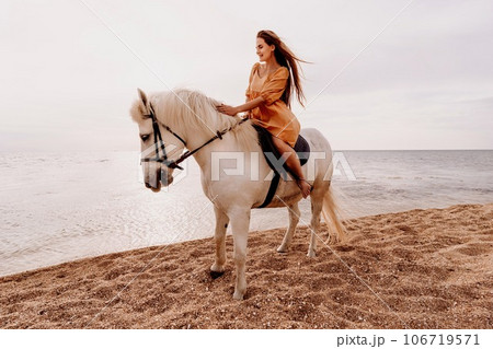 A white horse and a woman in a dress stand on a beach, with the sky and sea creating a picturesque backdrop for the scene. 106719571