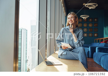 Smiling young woman freelancer sitting near window and making notes during working on laptop 106721392