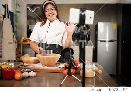 Young attractive Asian woman is blogging for her kitchen channel about homemade pizza while streaming online for webinar masterclass lesson. Vlogger broadcasting live video online teaching cooking Young attractive Asian woman is blogging for her kitchen channel about homemade pizza while streaming online for webinar masterclass lesson. Vlogger broadcasting live video online teaching cooking 106725194