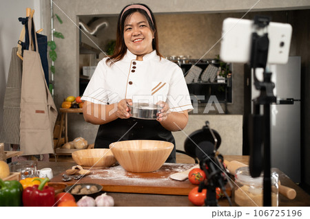 Young attractive Asian woman is blogging for her kitchen channel about homemade pizza while streaming online for webinar masterclass lesson. Vlogger broadcasting live video online teaching cooking Young attractive Asian woman is blogging for her kitchen channel about homemade pizza while streaming online for webinar masterclass lesson. Vlogger broadcasting live video online teaching cooking 106725196