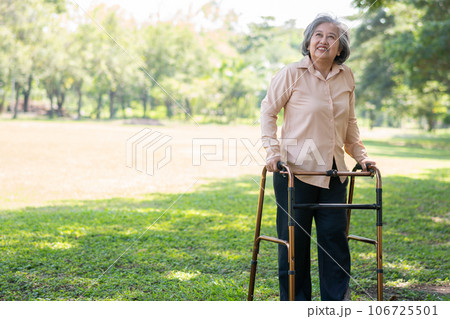 Happy old elderly Asian woman uses a walker for osteoarthritis rehabilitation physiotherapy in park.  Concept of happy retirement With care from a caregiver and Savings and senior health insurance 106725501