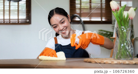 Happy young housewife in orange rubber gloves cleaning table countertop. Positive millennial maid keeping her home tidy and clean hygiene, wiping surface with microfiber cloth 106726360